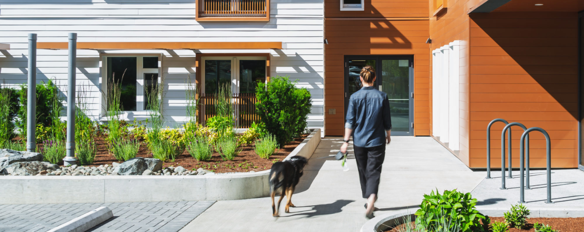 A woman walks her leashed dog head towards the entrance of a bright, modern apartment building
