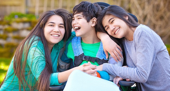 Image of three individuals sitting closely together outdoors. The individual in the center appears to be in a wheelchair, and the other two individuals are leaning in on either side, making physical contact with the central individual.